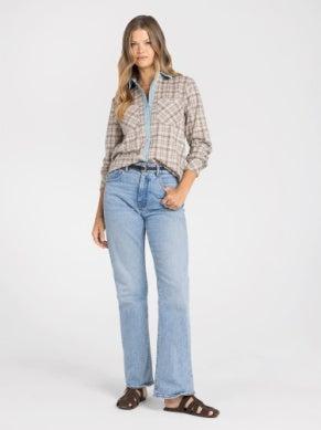 A woman modeling a brown and beige plaid button-down shirt with light-wash blue jeans and brown sandals, standing against a plain white studio background.