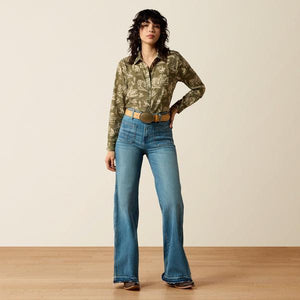 Woman wearing a patterned shirt and blue jeans standing against a plain background