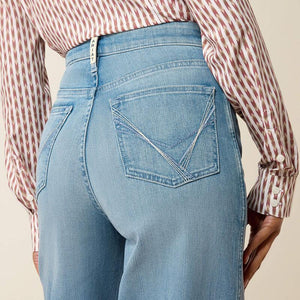 Close-up back view of a person wearing light-wash blue jeans with decorative back pocket stitching and a red-and-white striped button-down shirt, against a plain beige background.