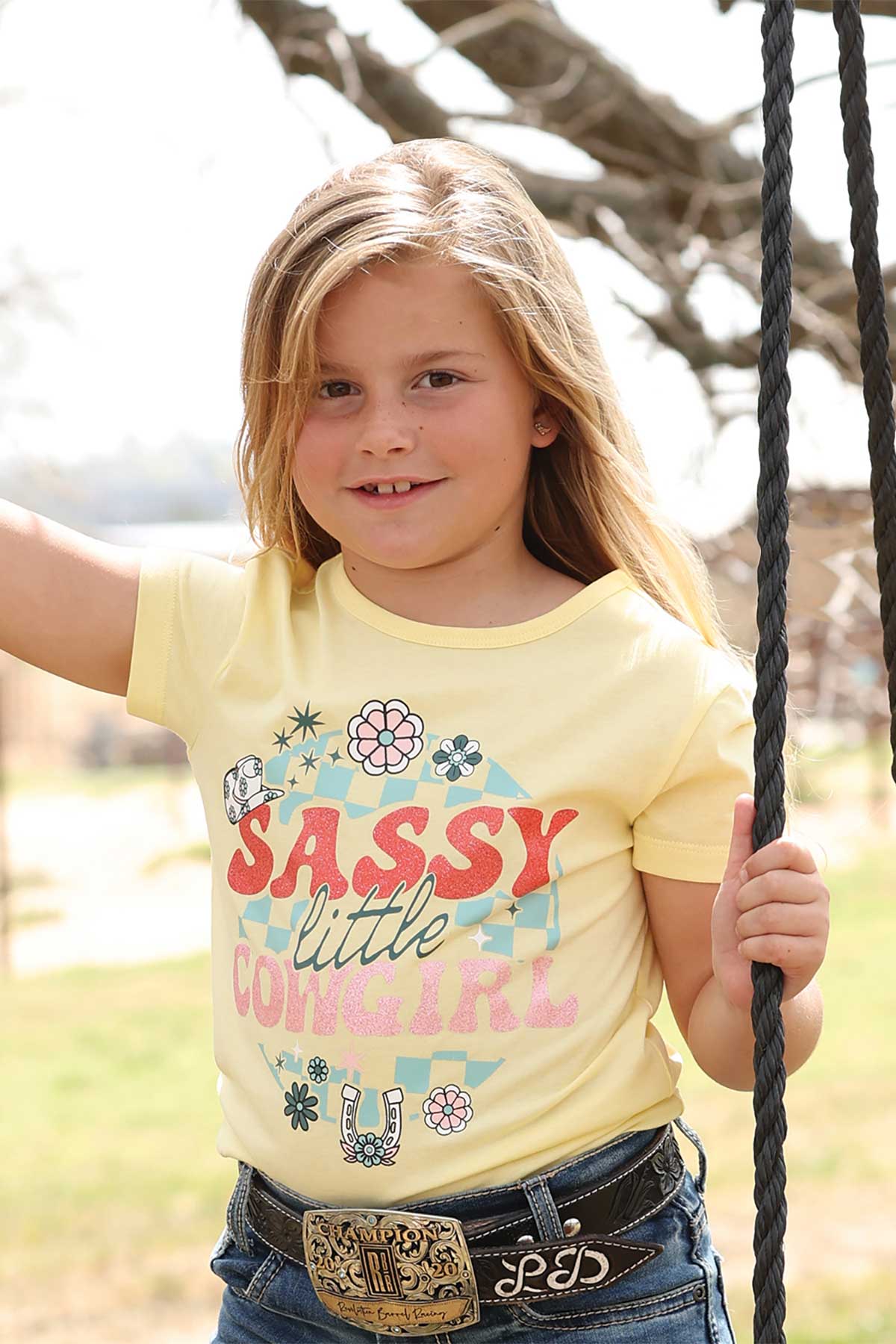 Young girl on a swing wearing a yellow t-shirt with 'Sassy little cowgirl' text.