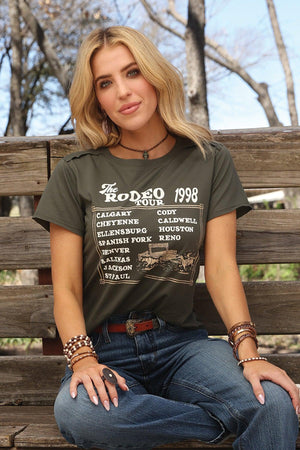 Woman wearing a t-shirt with 'The Rodeo 1998' design, sitting on a wooden bench outdoors.