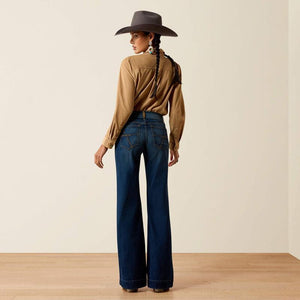 A model is shown from behind wearing a brown wide-brim hat, a tan long-sleeve button-up shirt, and dark blue wide-leg jeans, standing on a light wood floor against a beige wall.