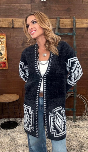 A woman wearing a long open-front knit cardigan with black and white Southwestern geometric patterns, styled with a white top and blue jeans in a rustic indoor setting.