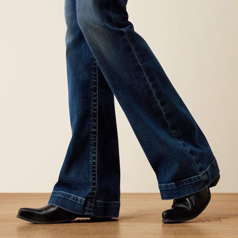 Close-up of a person's legs wearing dark blue flared jeans and black leather shoes, standing on a light wood floor against a plain beige background.