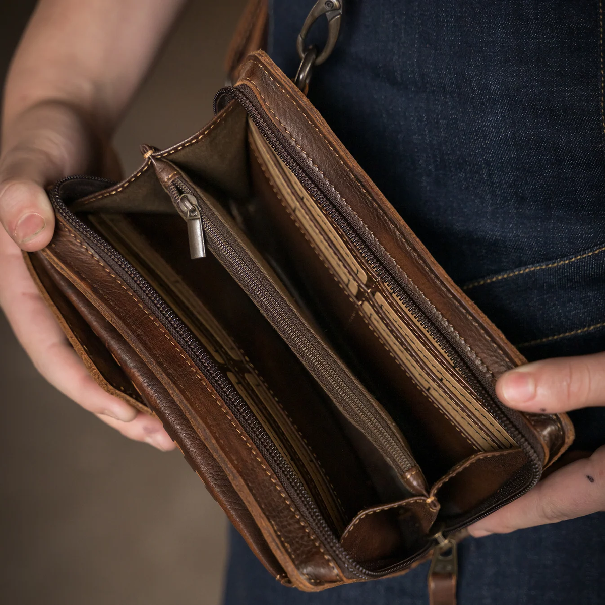 A close-up of a person holding an open brown leather wallet or clutch with multiple zip compartments and card slots, shown against dark denim clothing.
