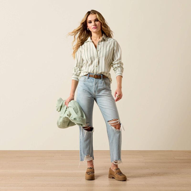 Woman wearing a striped shirt, light blue jeans, and brown boots holding a green jacket.