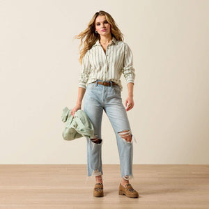 Woman wearing a striped shirt, light blue jeans, and brown boots holding a green jacket.