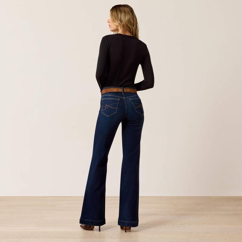 A woman is shown from the back wearing dark blue flare jeans, a black long-sleeve top, and brown heels, standing against a plain beige studio background.