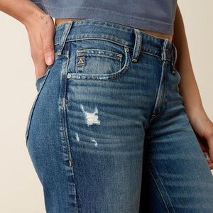 Close-up of a person wearing blue distressed denim jeans with a small gold 'A' logo on the pocket, paired with a gray cropped top against a light beige background.