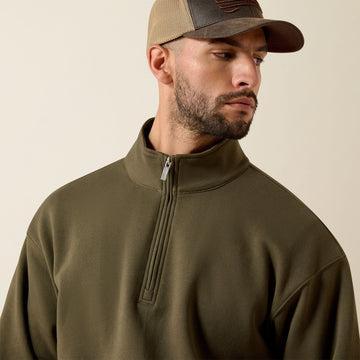 A man wearing an olive green quarter-zip fleece pullover and a brown and tan baseball cap, shown from the chest up against a plain beige background.