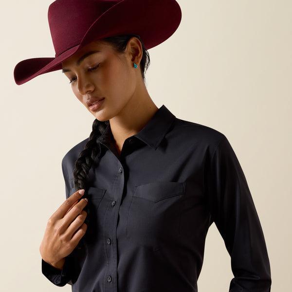 A woman wearing a black long-sleeve button-up shirt and a burgundy cowboy hat, looking down against a plain beige background.