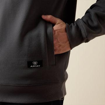 Close-up of a person wearing a dark charcoal gray hoodie, showing a hand in the side pocket and a small black Ariat logo patch on the lower front.
