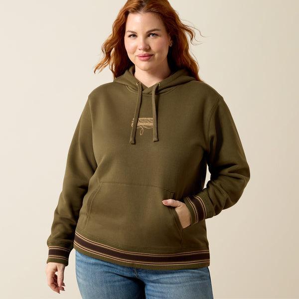 A woman wearing an olive green pullover hoodie with drawstrings, a kangaroo pocket, and ribbed cuffs and hem with thin tan stripes. 