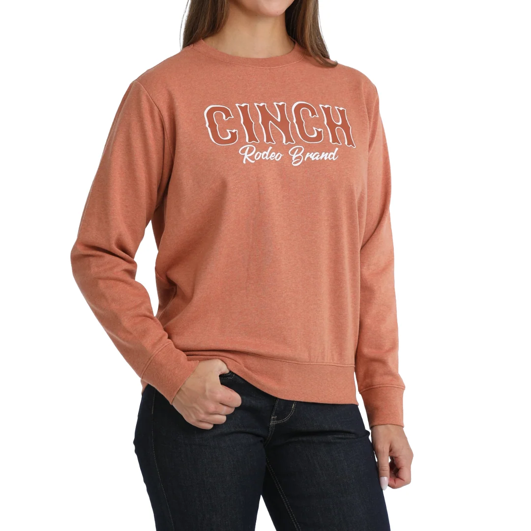 A woman wearing a heathered terracotta crewneck sweatshirt with 'CINCH Rideos Brand' printed in white and brown outline across the chest, paired with dark jeans, against a plain white background.