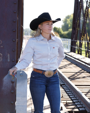 A woman wearing a black cowboy hat, a white button-down shirt with thin blue vertical stripes, and blue jeans stands on a bridge next to railroad tracks.