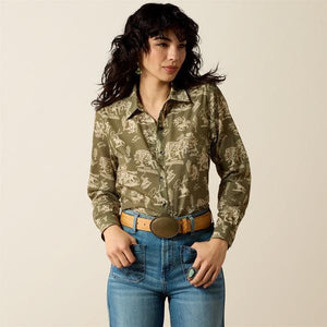 A woman modeling a long-sleeve button-up shirt with an olive green and beige western-style animal print, paired with blue jeans and a brown belt, against a solid light beige background.