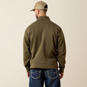 A man viewed from behind wearing an olive green quarter-zip fleece jacket, blue jeans, and a tan baseball cap against a plain beige background.