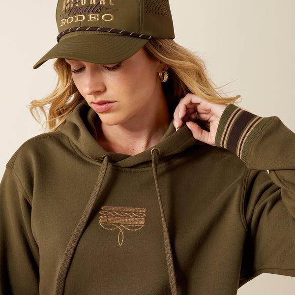 A woman wearing an olive green pullover hoodie with drawstrings, a kangaroo pocket, and ribbed cuffs and hem with thin tan stripes. 