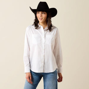 A woman wearing a white long-sleeve button-up shirt and a burgundy cowboy hat, looking down against a plain beige background.