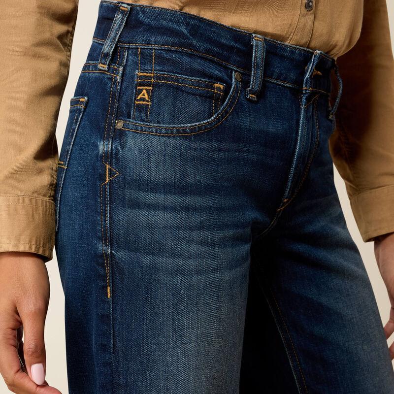 Close-up of a person wearing dark blue denim jeans with a tan shirt. The jeans show a small embroidered 'A' logo on the pocket and contrast gold stitching.