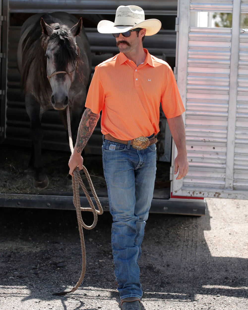 Man in orange shirt and cowboy hat holding a horse by a trailer