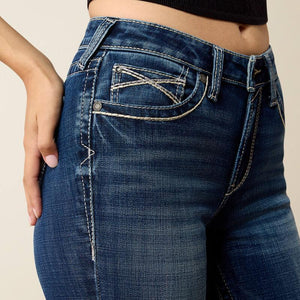 Close-up of a person wearing dark blue denim jeans with contrast light stitching and a decorative front pocket detail, paired with a black cropped top against a plain beige background.