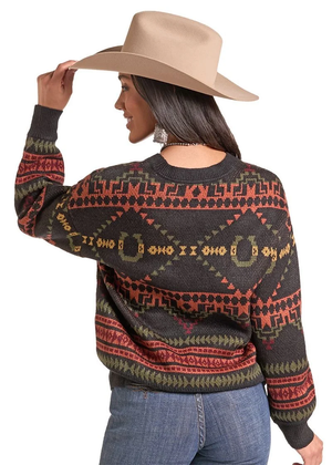 A woman wearing a dark charcoal knit sweater with geometric and horseshoe patterns in rust red, olive green, and tan, paired with a beige cowboy hat and blue jeans, shown from a three-quarters back view against a white background.