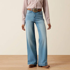A person wearing light blue wide-leg denim jeans with a brown belt and a pink-and-white striped shirt, standing against a plain beige wall on a light wood floor.