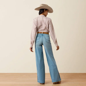 A woman is shown from behind wearing a light beige cowboy hat, a pink and white patterned long-sleeve cropped shirt, and light blue wide-leg jeans, standing on a light wood floor against a beige wall.