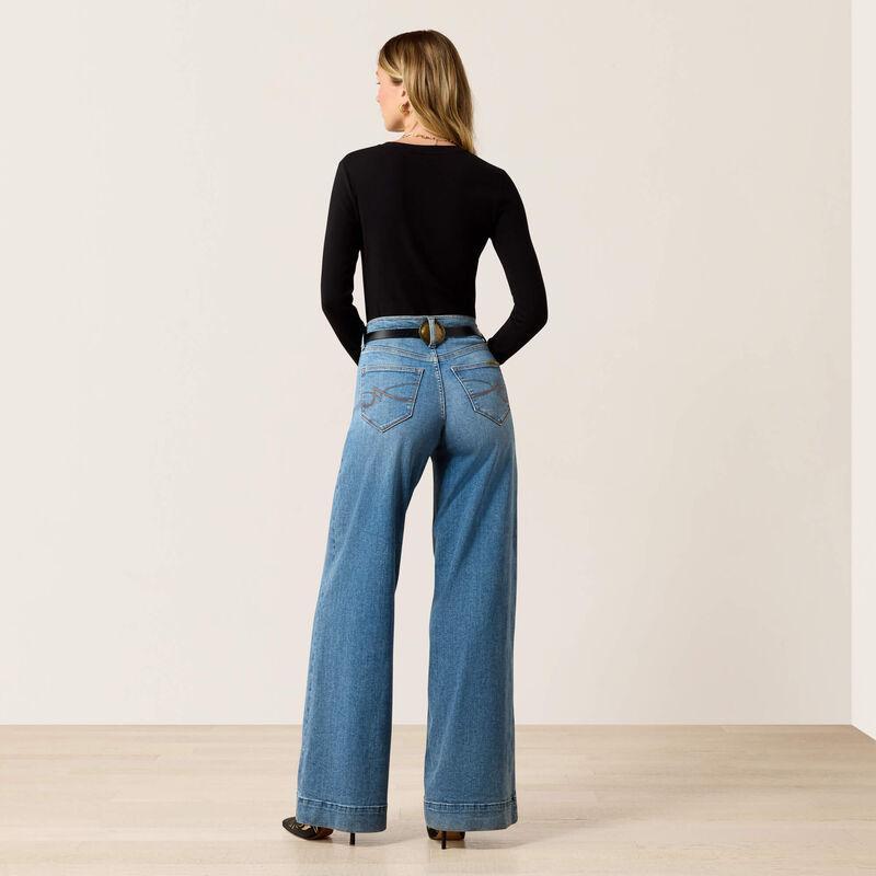 A woman stands with her back to the camera wearing a fitted black long-sleeve top, wide-leg blue jeans, and black heels against a plain beige studio wall and light wood floor.