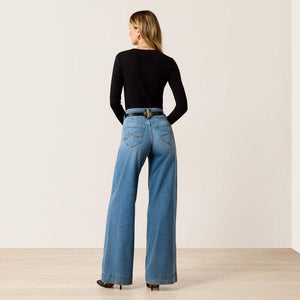 A woman stands with her back to the camera wearing a fitted black long-sleeve top, wide-leg blue jeans, and black heels against a plain beige studio wall and light wood floor.