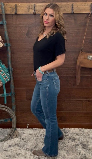 Woman wearing a black top and blue jeans standing against a wooden wall with rustic decor.