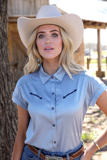 Woman wearing a light blue shirt with black arrows and a beige cowboy hat outdoors.
