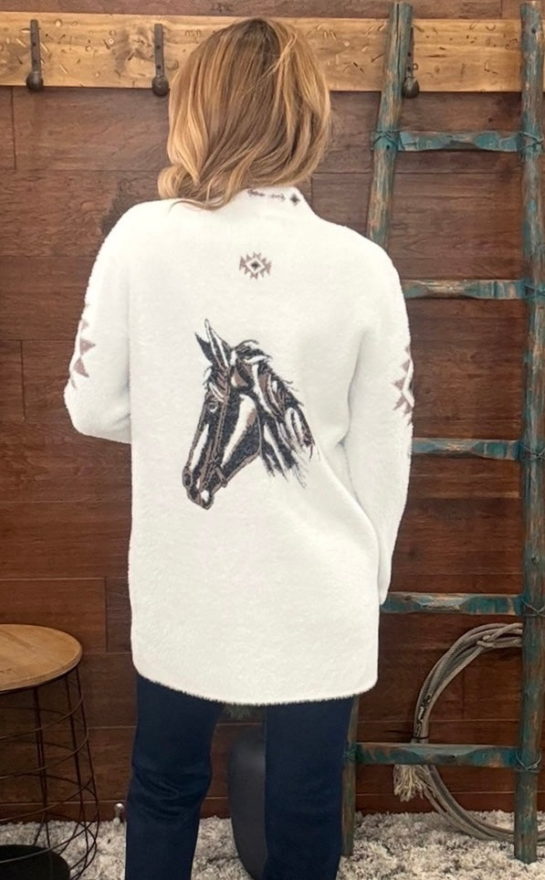 A woman viewed from behind wearing a cream-colored knit sweater with a large black horse graphic on the back, paired with dark blue jeans, standing in front of a rustic wooden wall with a ladder.