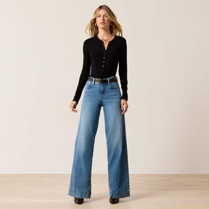 A woman modeling a black long-sleeve henley top and high-waisted, wide-leg blue jeans with a black belt, standing against a neutral beige studio background.