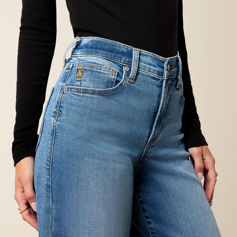 Close-up of a person wearing high-rise blue denim jeans with a black long-sleeve top, shown from the chest down against a plain light beige background.