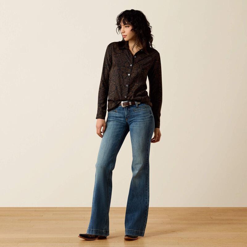 A woman stands in a studio wearing a dark brown patterned button-up shirt and blue flare jeans, paired with a brown belt and dark brown shoes, against a warm beige background.