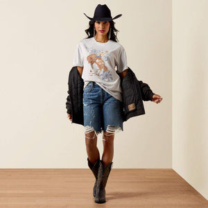 Woman wearing a white t-shirt with a graphic design, black jacket, denim shorts, and cowboy boots on a beige background.