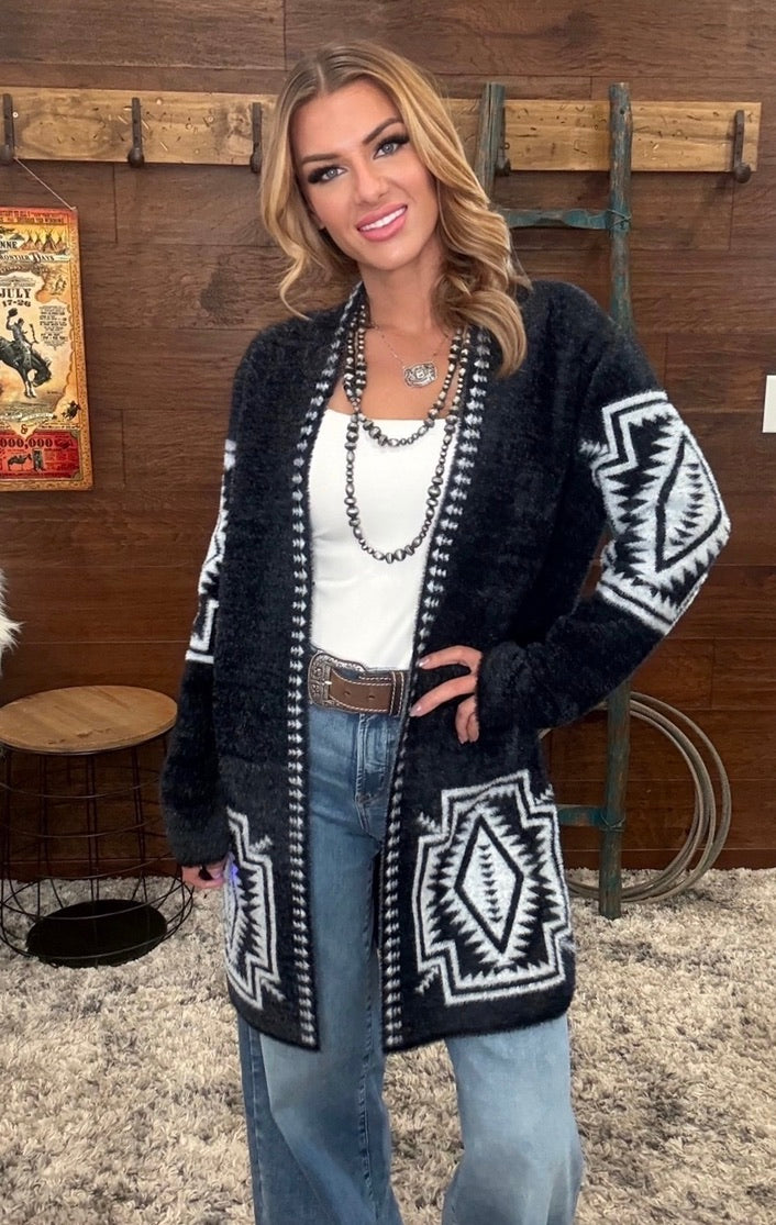 A woman modeling a black fuzzy open-front cardigan with white geometric Southwestern-style patterns on the sleeves and lower front, styled over a white top and blue jeans in a rustic indoor setting.