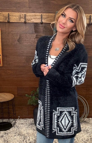 A woman wearing a black fuzzy cardigan with white geometric patterns, standing in front of a rustic wood-paneled wall.