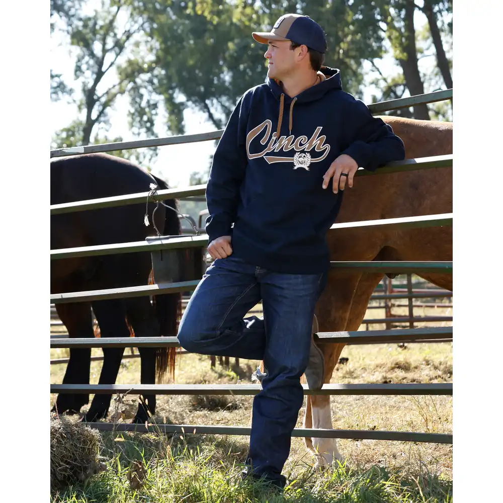 A man wearing a navy blue hoodie with 'Cinch' written in white script across the chest, blue jeans, and a baseball cap, leaning against a metal fence in a ranch setting with horses behind him.