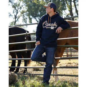 A man wearing a navy blue hoodie with 'Cinch' written in white script across the chest, blue jeans, and a baseball cap, leaning against a metal fence in a ranch setting with horses behind him.
